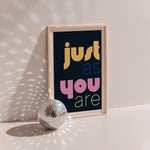 Just As You Are poster in wood frame beside a disco ball, multicoloured lettering on black