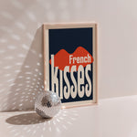 French Kisses poster in wood frame beside a disco ball, red lips with cream warped lettering on navy