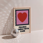 Fragile Handle With Care poster in wood frame beside a disco ball, postage stamp frame with red heart on pink