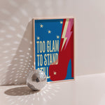 Too Glam poster in wood frame beside a disco ball, red platform boot with lightning bolt on blue