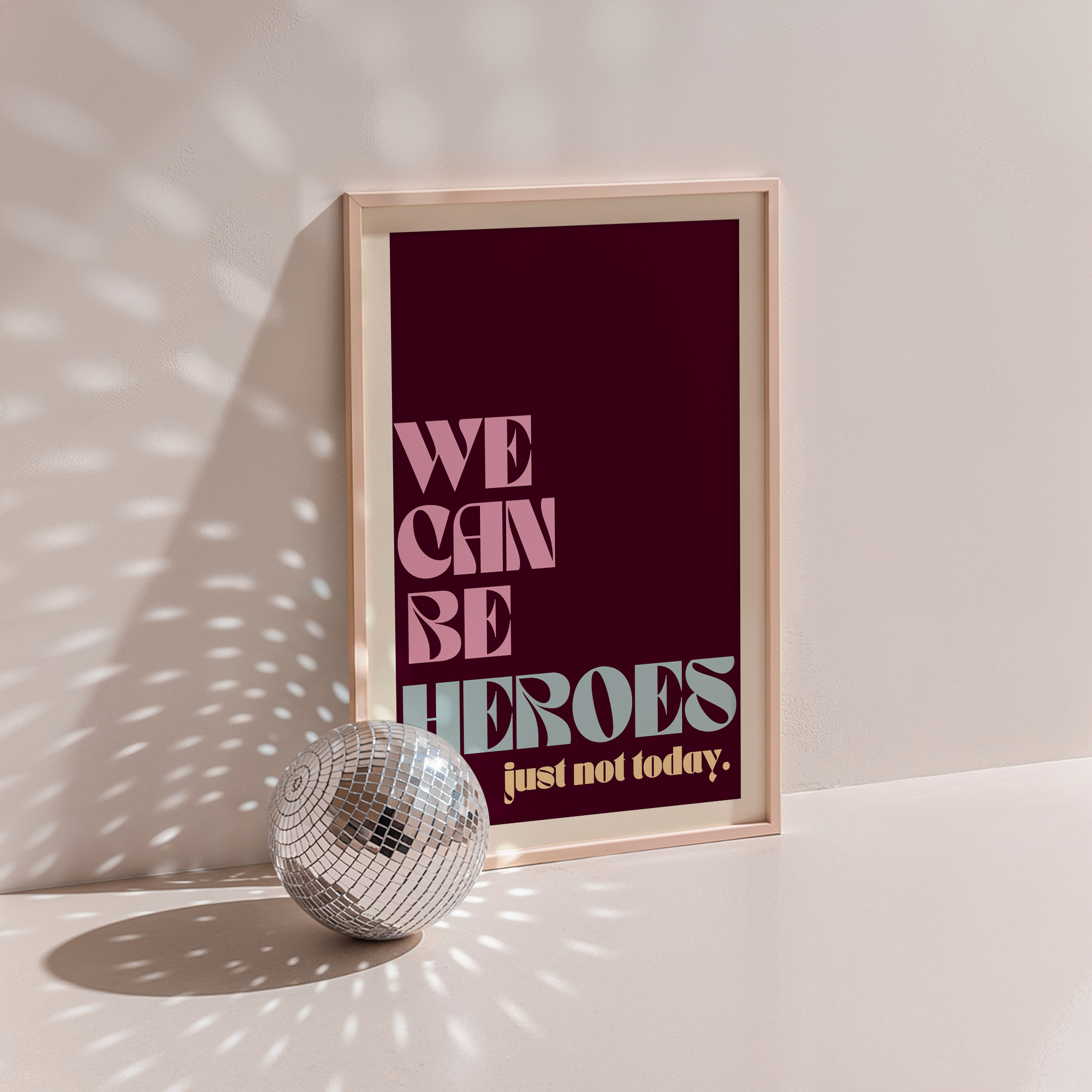 We Can Be Heroes poster in wood frame beside a disco ball, burgundy background with cream lettering and 'just not today' subline